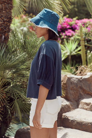 Claudia tee in navy with Natalie shorts in ecru and denim fringed bucket hat, with model standing to the side to show puff sleeve.